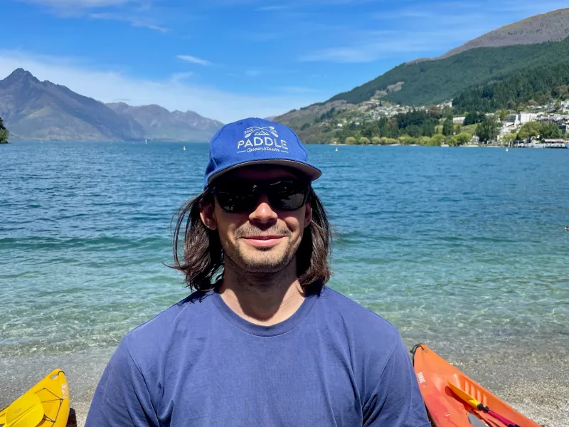 Person in blue shirt and cap stands by lake with mountains and kayaks in background.